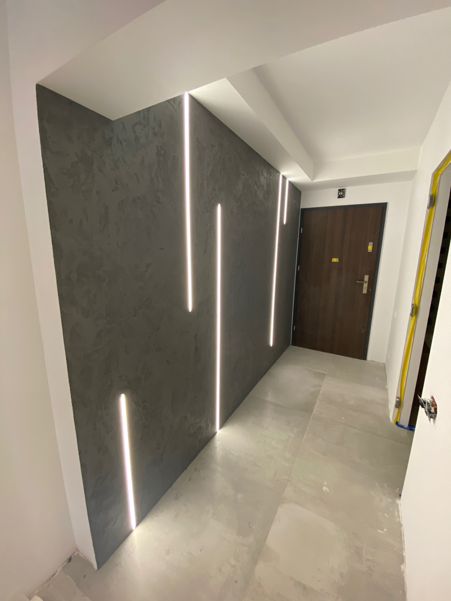 Korytarz z szarą ścianą o fakturze betonu, oświetlony pionowymi paskami LED, z widoczną brązową ścianą i szarą podłogą z płytek, poziomica oparta o ścianę.