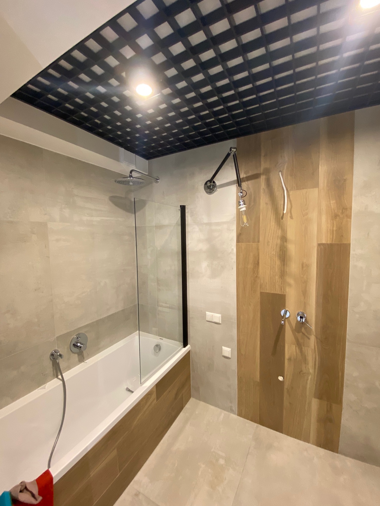 Nowoczesna łazienka z wanną z parawanem, drewnopodobnymi płytkami na ścianie i sufitem z czarną kratką, oświetlona designerską lampą z żarówką Edisona.