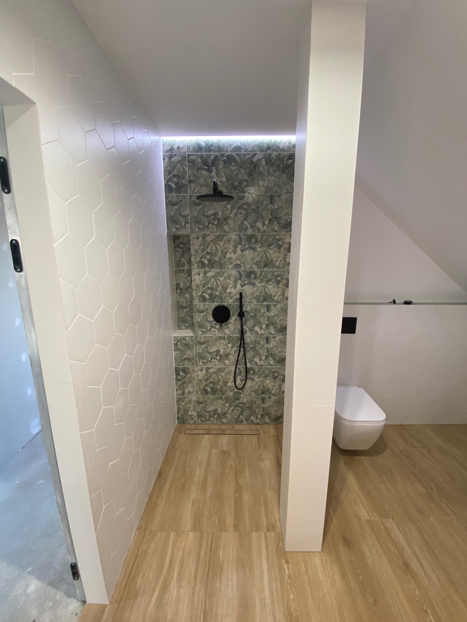 Nowoczesna łazienka na poddaszu z kabiną prysznicową typu walk-in, heksagonalnymi płytkami na ścianach, panelami z motywem roślinnym w strefie prysznica, drewnopodobną podłogą i podświetleniem LED