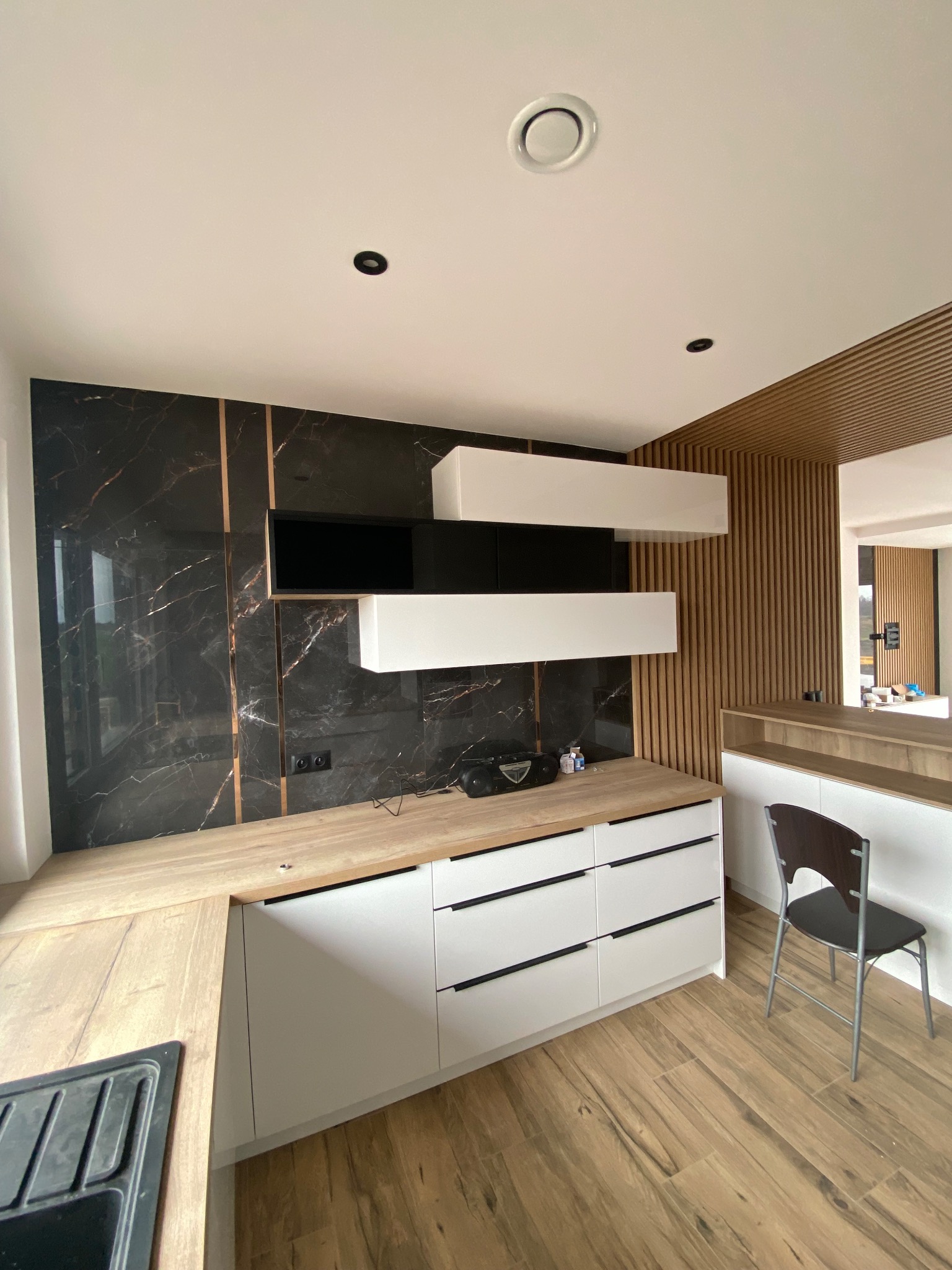 Nowoczesna kuchnia z czarnym marmurem na ścianie, drewnianymi lamelami na suficie i ścianie oraz białymi szafkami z drewnianym blatem, uzupełniona drewnianą podłogą i czarnym krzesłem.