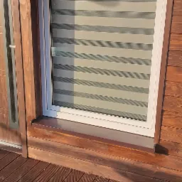 Białe okno z żaluzjami poziomymi w drewnianej elewacji domu, widok z zewnątrz na drewniany taras.