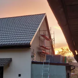 Dom jednorodzinny w trakcie prac elewacyjnych przy zachodzie słońca, widoczne rusztowanie, drabina oparta o ścianę i materiały budowlane na dachu dobudówki.