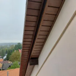 Wykończone drewnianą boazerią podbitka dachowa i fragment elewacji budynku z widokiem na okoliczne domy w pochmurny dzień.