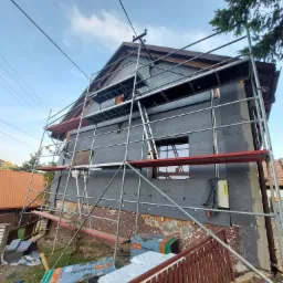 Dom w trakcie ocieplania styropianem grafitowym, widoczne rusztowanie, częściowo otynkowana dolna partia elewacji, opakowania styropianu Termo Organika Galaxy leżące przy ogrodzeniu.