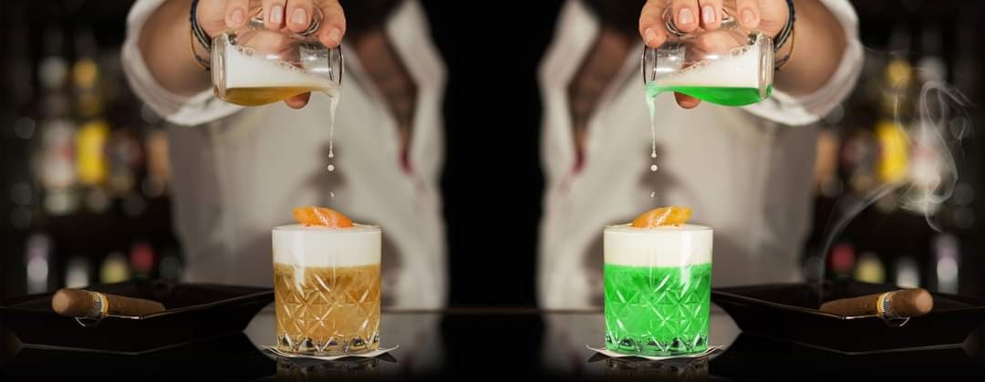 Barman nalewa płyn do dwóch koktajli z pianką i pomarańczą, obok leżą cygara, w tle butelki alkoholu.