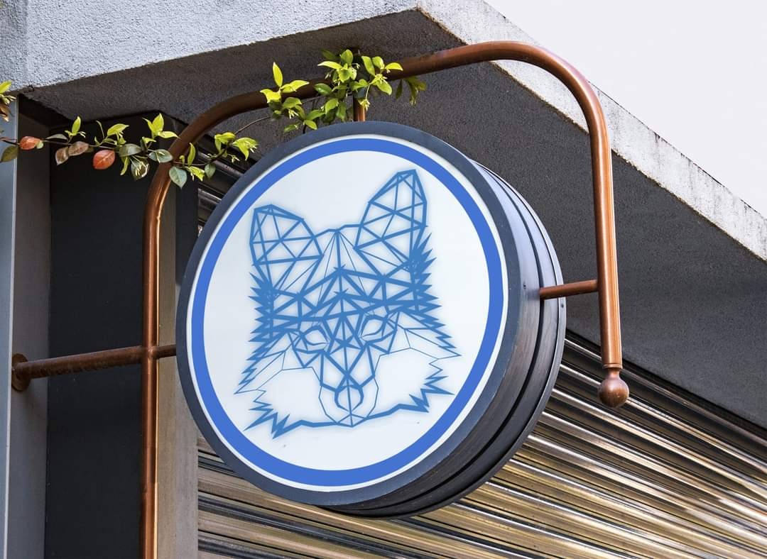 Okrągły szyld z geometrycznym wizerunkiem wilka w kolorze niebieskim, przymocowany do budynku w Warszawie za pomocą metalowej konstrukcji, z fragmentem zieleni w tle.