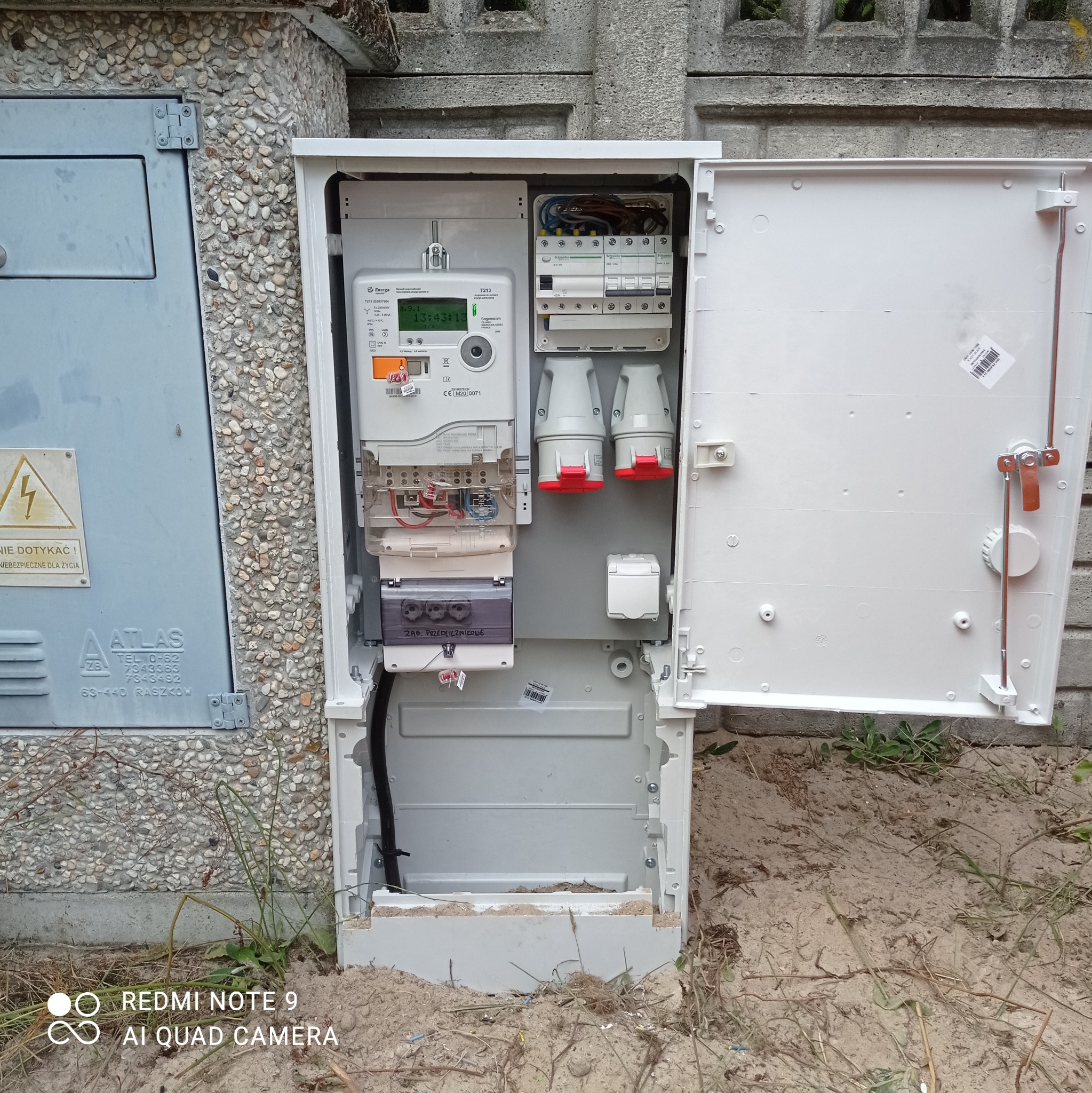 Otwarta skrzynka elektryczna z licznikiem i zabezpieczeniami, zamontowana na zewnątrz budynku, widoczne przewody i elementy instalacji, obok szara skrzynka z ostrzeżeniem o wysokim napięciu.