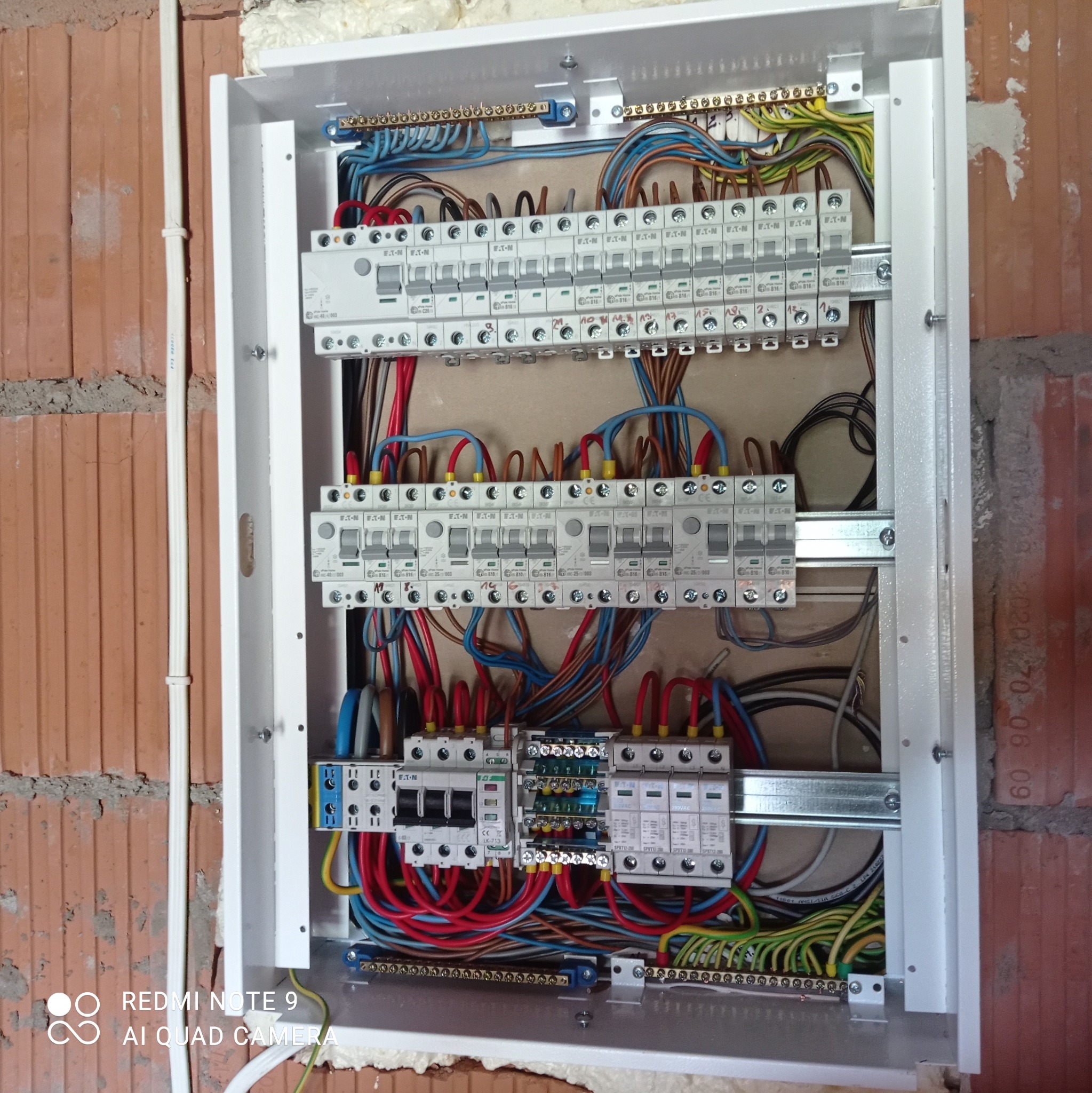 Otwarta skrzynka rozdzielcza z widocznymi bezpiecznikami, przewodami w różnych kolorach i listwami zaciskowymi, zamontowana na ścianie z cegły.