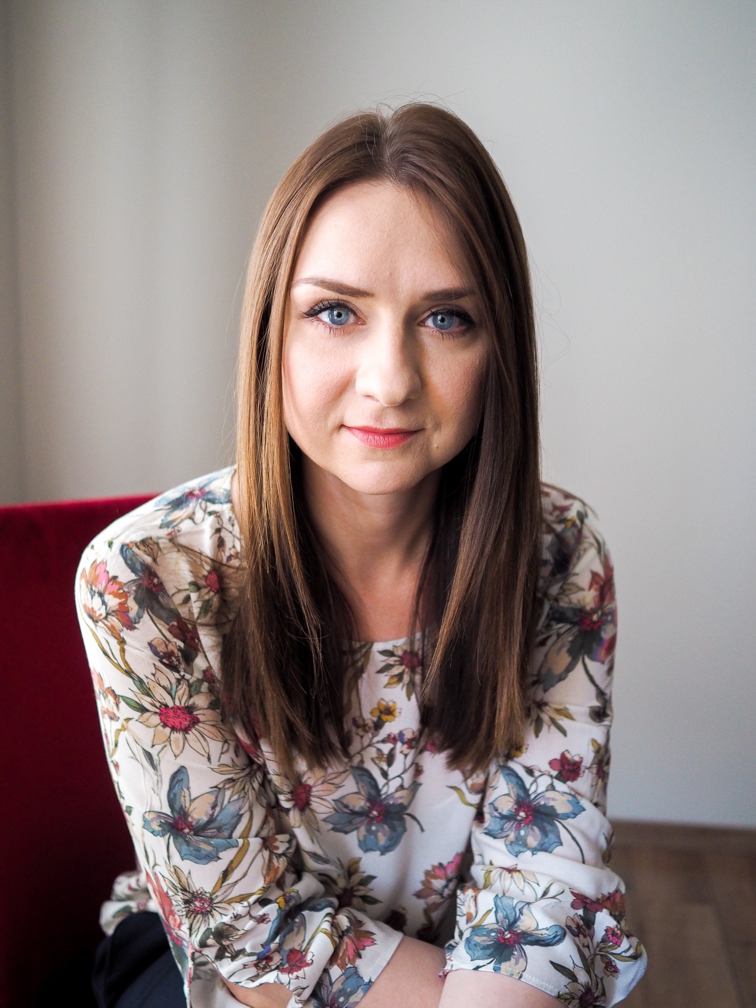 Portret kobiety z długimi brązowymi włosami i niebieskimi oczami, ubrana w kwiecistą bluzkę, siedząca na czerwonym krześle na tle jasnoszarej ściany.