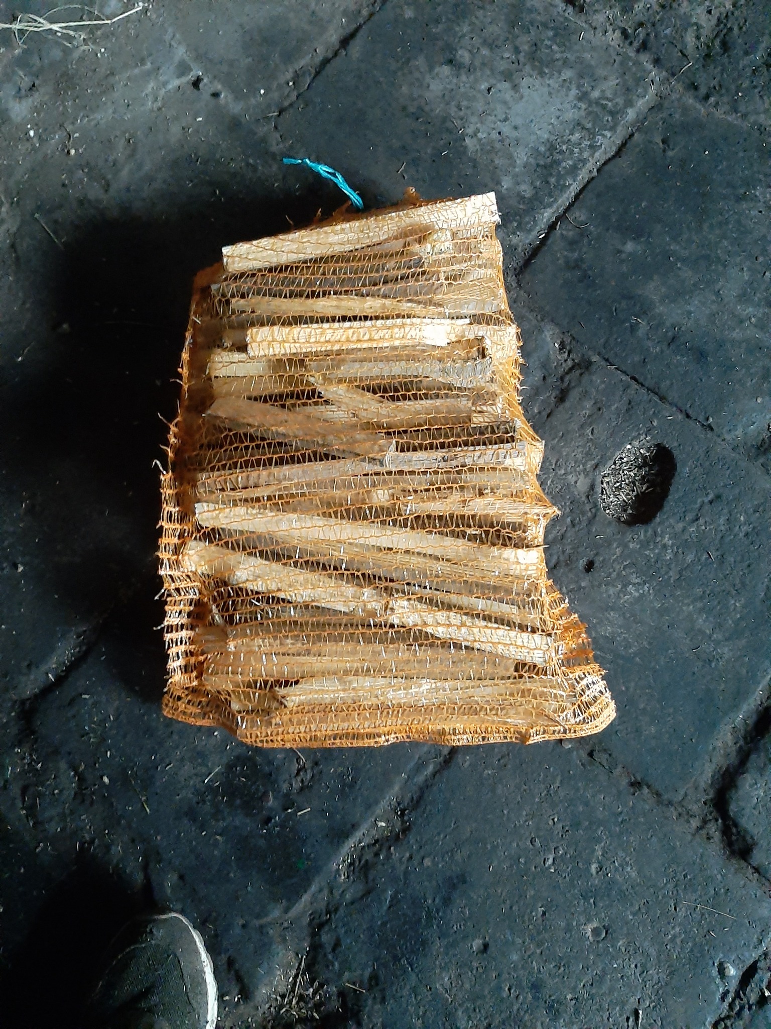 Worek z pociętym drewnem kominkowym, zamknięty niebieskim sznurkiem, leżący na ciemnej, popękanej posadzce.