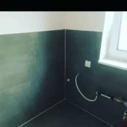 Wykończona łazienka z szarymi płytkami na ścianach i podłodze, widoczna instalacja wodna przy oknie.