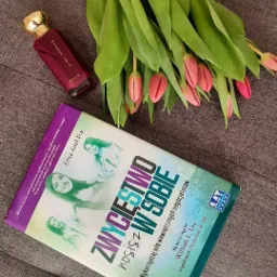 Książka 'Zwycięstwo w Sobie' autorstwa Erica Ariel Fox obok flakonu perfum Magnétista Oriflame i bukietu różowych tulipanów na szarej tkaninie.