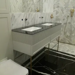 Nowoczesna łazienka z podwójną umywalką na szafce z pionowymi żłobieniami, czarnym blatem i złotymi nóżkami, marmurowe ściany w odcieniach bieli i szarości, wzorzysta podłoga.