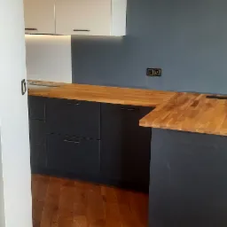 Nowoczesna kuchnia z drewnianym blatem i ciemnymi szafkami dolnymi, białe szafki wiszące z podświetleniem, drewniana podłoga i szara ściana z gniazdkami.