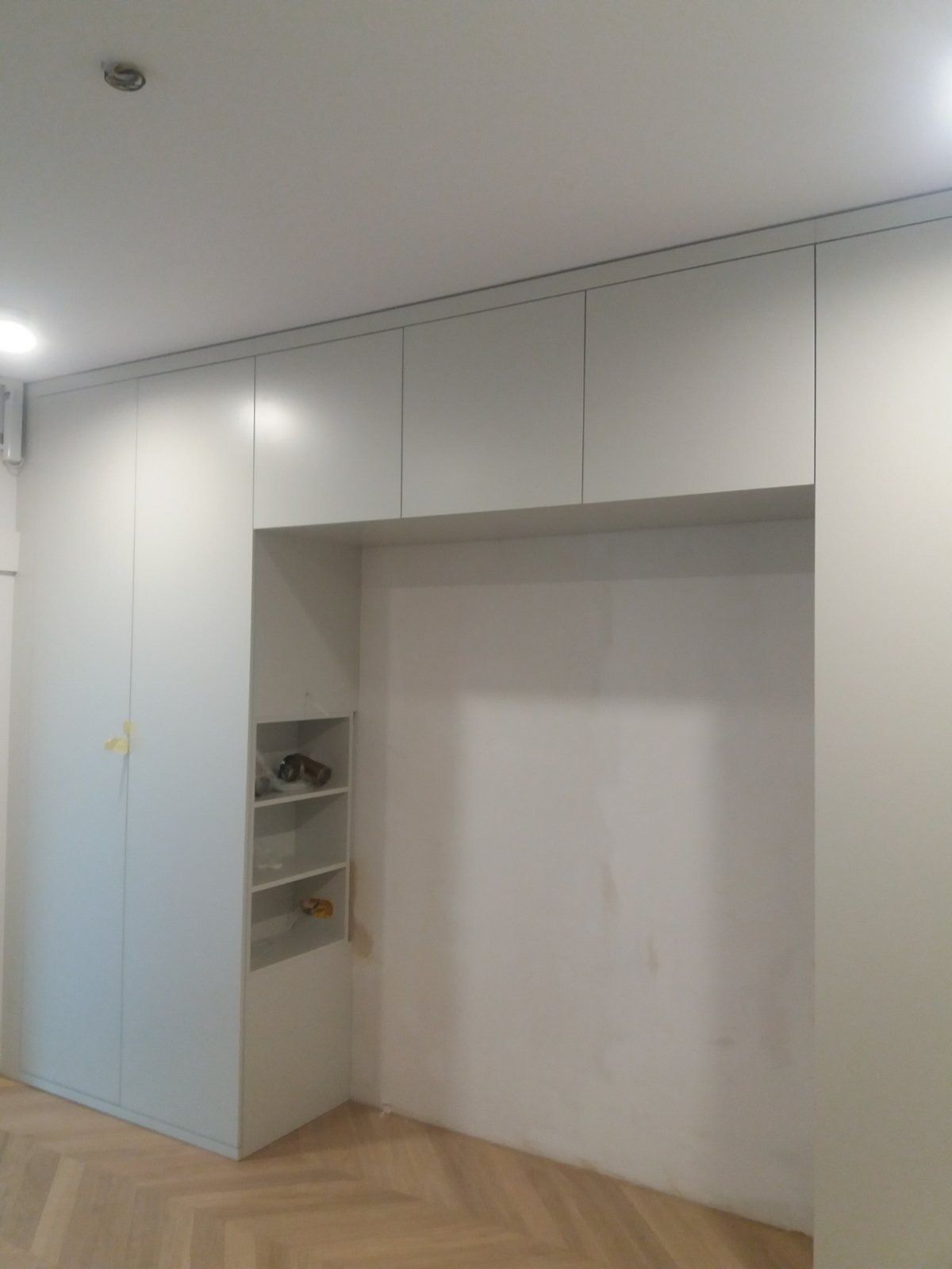 Jasnoszara zabudowa meblowa z otwartą wnęką z półkami, widoczna podłoga parkietowa w jodełkę, ściany przygotowane do malowania.