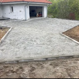 Przygotowany podjazd z obrzeżami przed domem z otwartym garażem, teren częściowo wyrównany, widoczne ślady opon na ziemi.