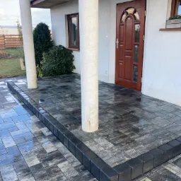 Nowo ułożony, ciemnoszary chodnik z kostki brukowej przed wejściem do domu z drewnianymi drzwiami, widoczne dwa białe filary i fragment ogrodu.