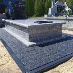 Szary nagrobek z ciemną tablicą i wizerunkiem Matki Boskiej, otoczony kostką brukową, na piaszczystym podłożu, w tle zielone drzewa.