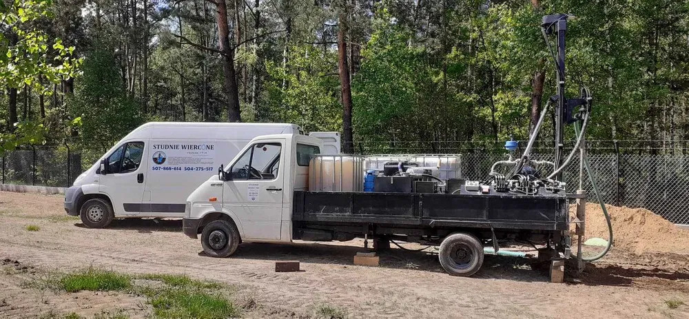 Wiertnica zamontowana na samochodzie ciężarowym podczas pracy na zewnątrz, obok zaparkowany bus z napisem 'Studnie Wiercone', widok na las w tle.