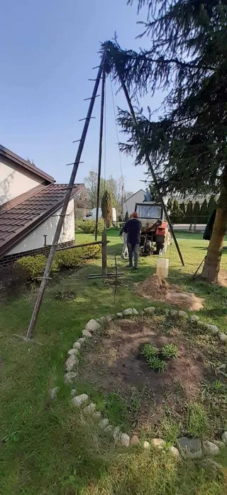 Montaż studni abisyńskiej na działce: stalowa konstrukcja w kształcie litery A, traktor w tle, pracownik w trakcie wiercenia, widoczny fragment domu.