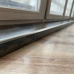 VDA REMONTY SPÓŁKA Z OGRANICZONĄ ODPOWIEDZIALNOŚCIĄ - Ciemna, kamienna parapetówka okienna z metalowym wykończeniem, zamontowana w jasnej ramie okiennej, sąsiadująca z panelami podłogowymi imitującymi drewno.