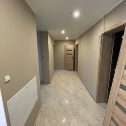 VDA REMONTY SPÓŁKA Z OGRANICZONĄ ODPOWIEDZIALNOŚCIĄ - Korytarz w mieszkaniu po odświeżeniu, beżowe ściany, marmurowe płytki na podłodze, widoczne drzwi z drewnianymi okleinami i niedokończone wykończenie ościeżnic.