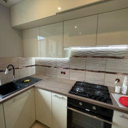 VDA REMONTY SPÓŁKA Z OGRANICZONĄ ODPOWIEDZIALNOŚCIĄ - Jasna kuchnia z beżowymi szafkami, czarnym zlewem i płytą gazową, ozdobne płytki ścienne z falistym wzorem i brązowymi akcentami.