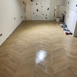 VDA REMONTY SPÓŁKA Z OGRANICZONĄ ODPOWIEDZIALNOŚCIĄ - Ukończona podłoga z płytek imitujących drewno ułożonych w jodełkę w łazience. Częściowo wykończone ściany z widocznymi puszkami elektrycznymi i płytkami dekoracyjnymi.