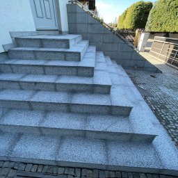 VDA REMONTY SPÓŁKA Z OGRANICZONĄ ODPOWIEDZIALNOŚCIĄ - Granitowe schody wejściowe z metalową barierką, prowadzące do białych drzwi. Tło stanowią zielone krzewy i kostka brukowa.