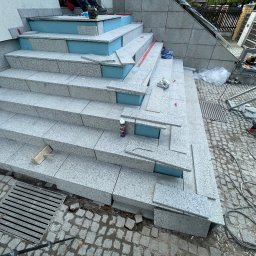 VDA REMONTY SPÓŁKA Z OGRANICZONĄ ODPOWIEDZIALNOŚCIĄ - Montaż granitowych płytek na schodach zewnętrznych. Widoczne narzędzia glazurnicze, poziomica, klej. Etap prac: płytki częściowo ułożone, z niebieską izolacją.