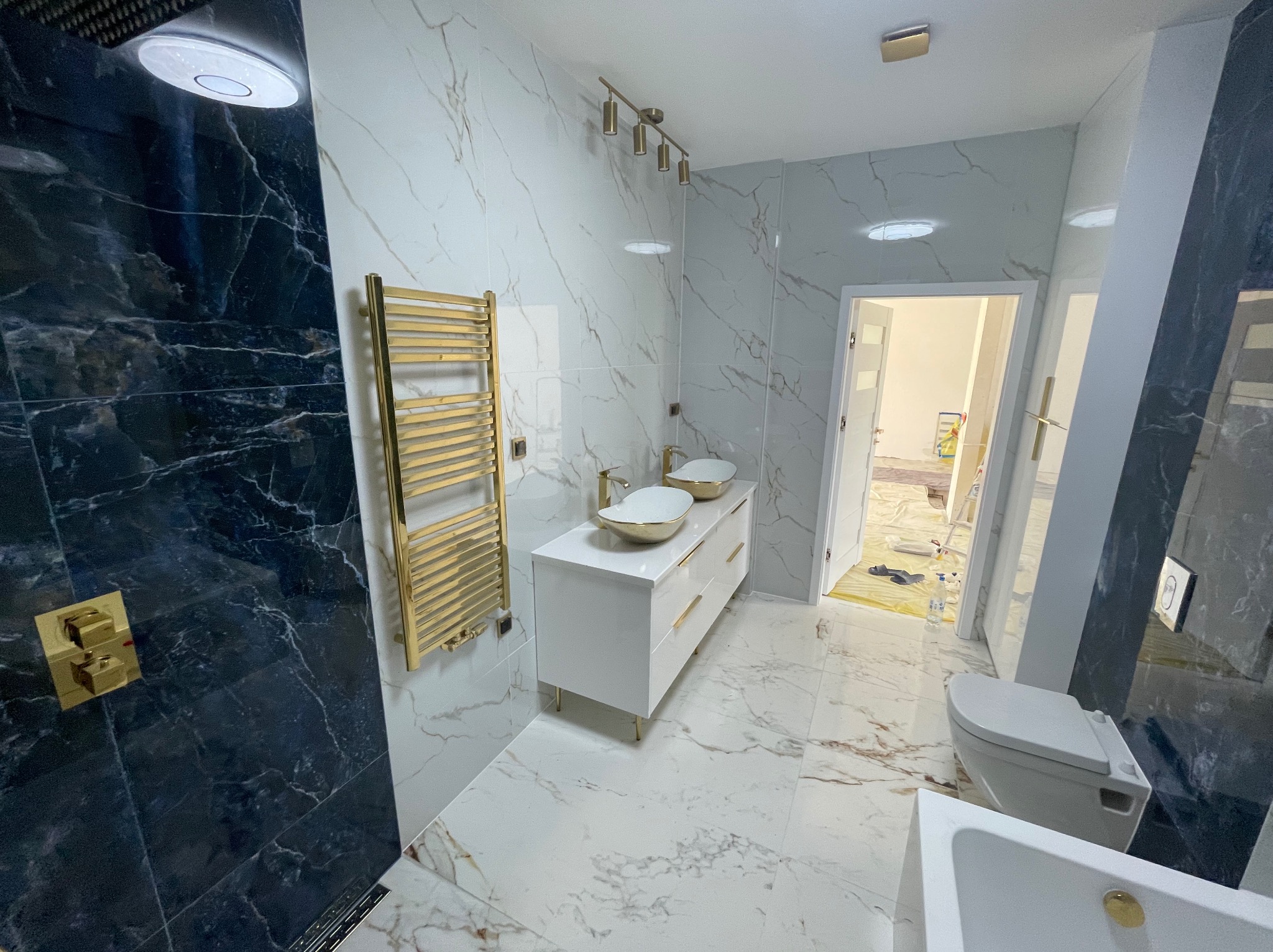 Nowoczesna łazienka z czarnymi marmurowymi płytkami w strefie prysznica, białymi marmurowymi płytkami na pozostałych ścianach, złotym grzejnikiem drabinkowym i podwójną umywalką nablatową na białej...