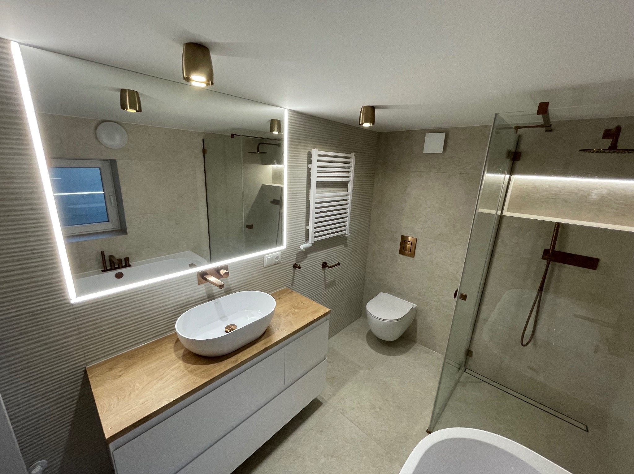 Nowoczesna łazienka z owalną umywalką nablatową na drewnianym blacie, podświetlanym lustrem, grzejnikiem drabinkowym i kabiną prysznicową z deszczownicą w kolorze miedzi.