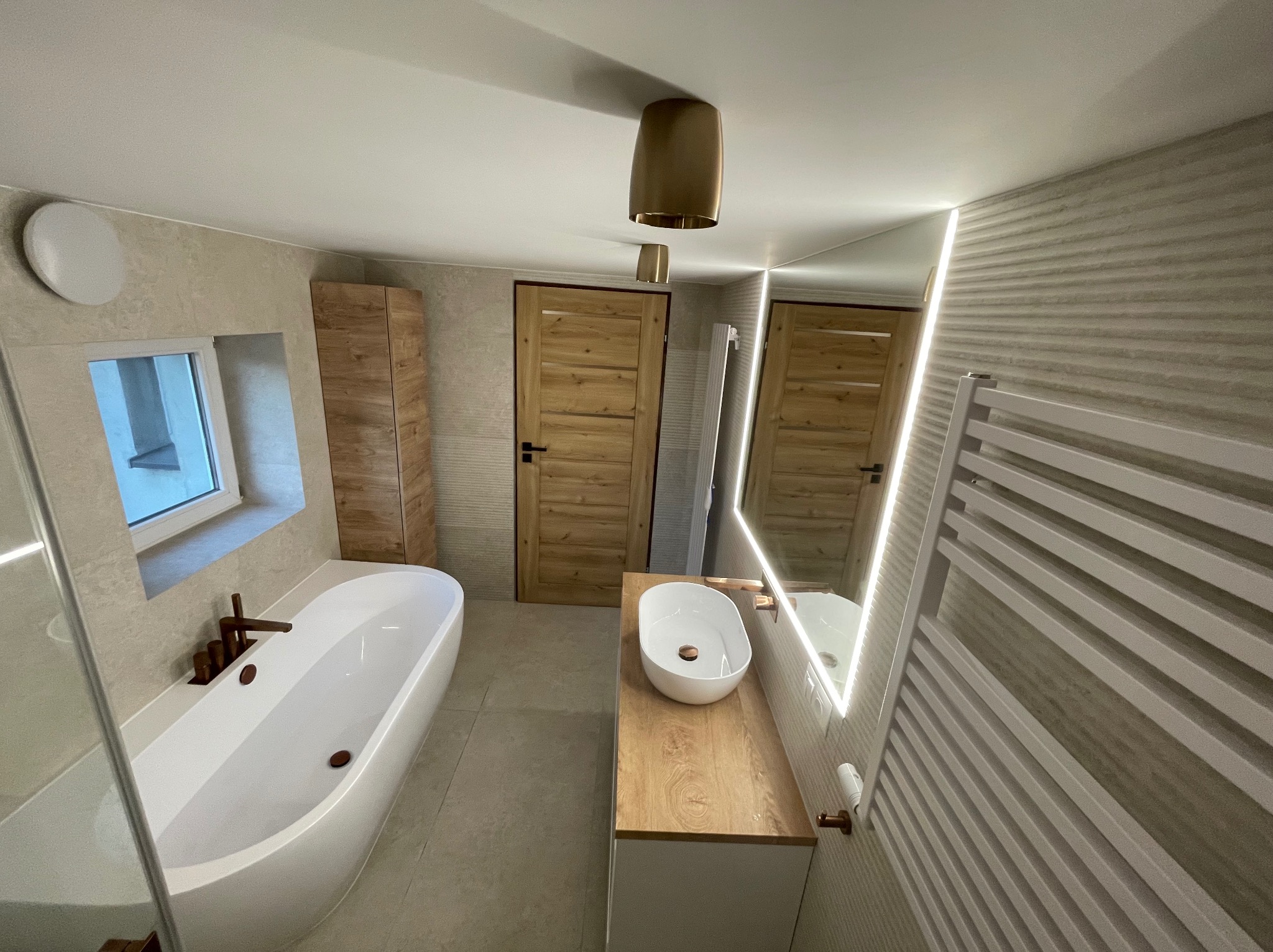 Nowoczesna łazienka z wolnostojącą wanną, umywalką nablatową na drewnianym blacie, podświetlanym lustrem LED i grzejnikiem drabinkowym, wykończona w beżowej kolorystyce z drewnianymi akcentami.