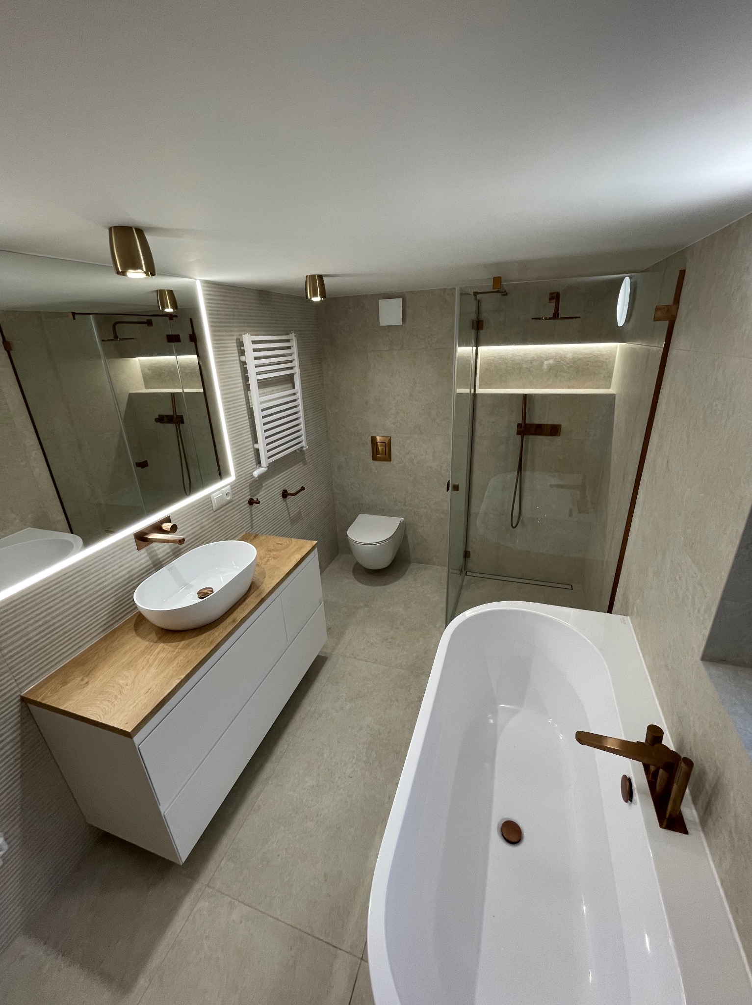 Nowoczesna łazienka w beżowych tonach z drewnianym blatem pod umywalką, kabiną prysznicową typu walk-in i wanną z miedzianą armaturą.