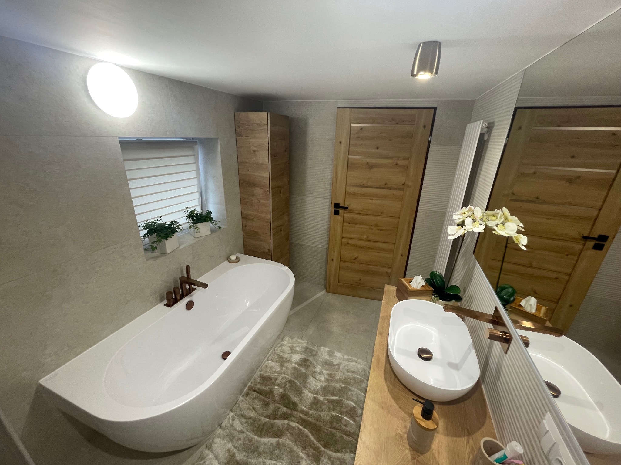 Nowoczesna łazienka z wolnostojącą wanną, umywalką nablatową i drewnianymi akcentami, oświetlona okrągłą lampą sufitową i z roślinami w oknie.