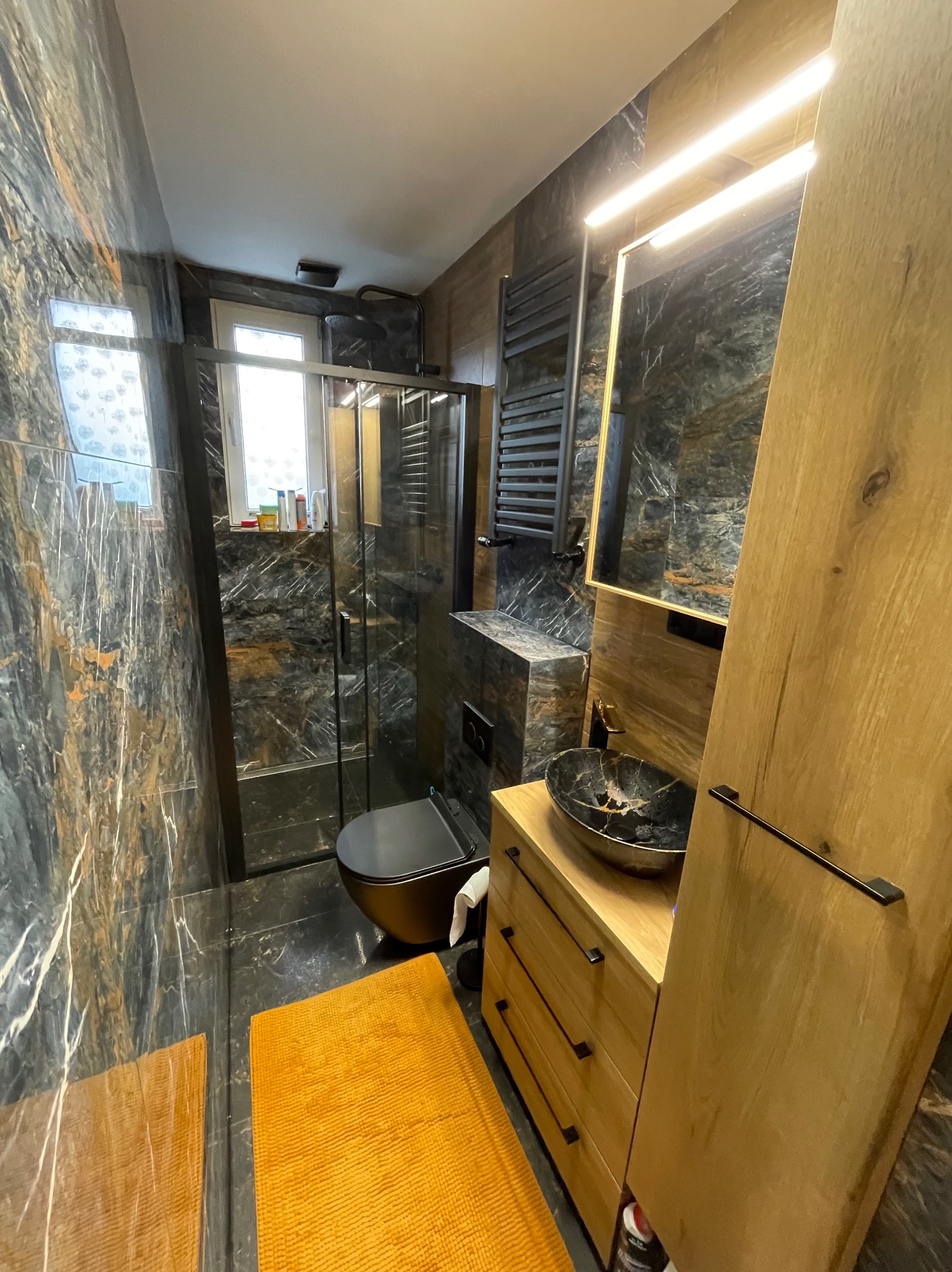 Nowoczesna łazienka z czarnymi marmurowymi płytkami, kabiną prysznicową, umywalką w stylu kamiennym i drewnianymi szafkami. Żółty dywanik dodaje akcentu kolorystycznego.