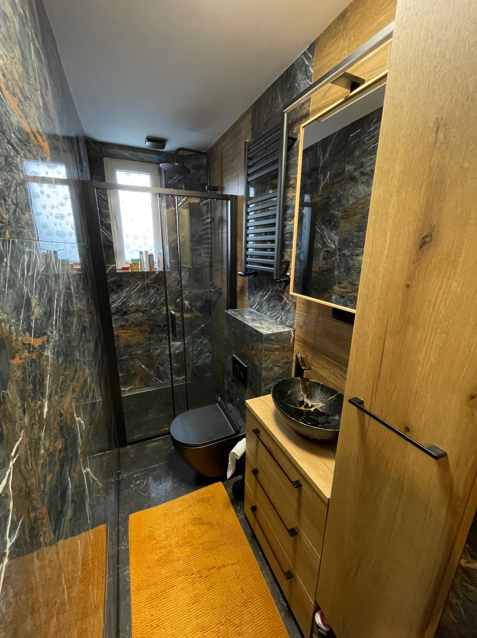 Wykończona łazienka z kabiną prysznicową, umywalką w kolorze czarnym i grzejnikiem drabinkowym. Marmurowe płytki na ścianach i podłodze. Nowoczesny design.