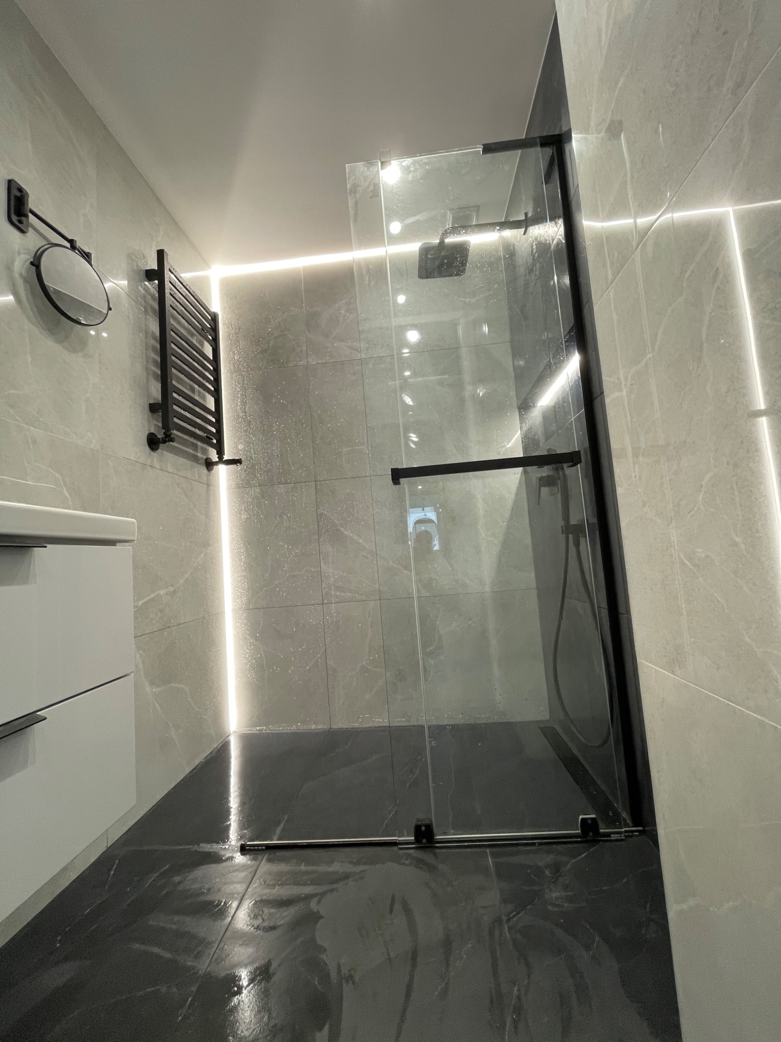 Nowoczesna łazienka z kabiną prysznicową typu walk-in, czarną armaturą i grzejnikiem drabinkowym. Ściany i podłoga wyłożone płytkami imitującymi marmur, oświetlenie LED.