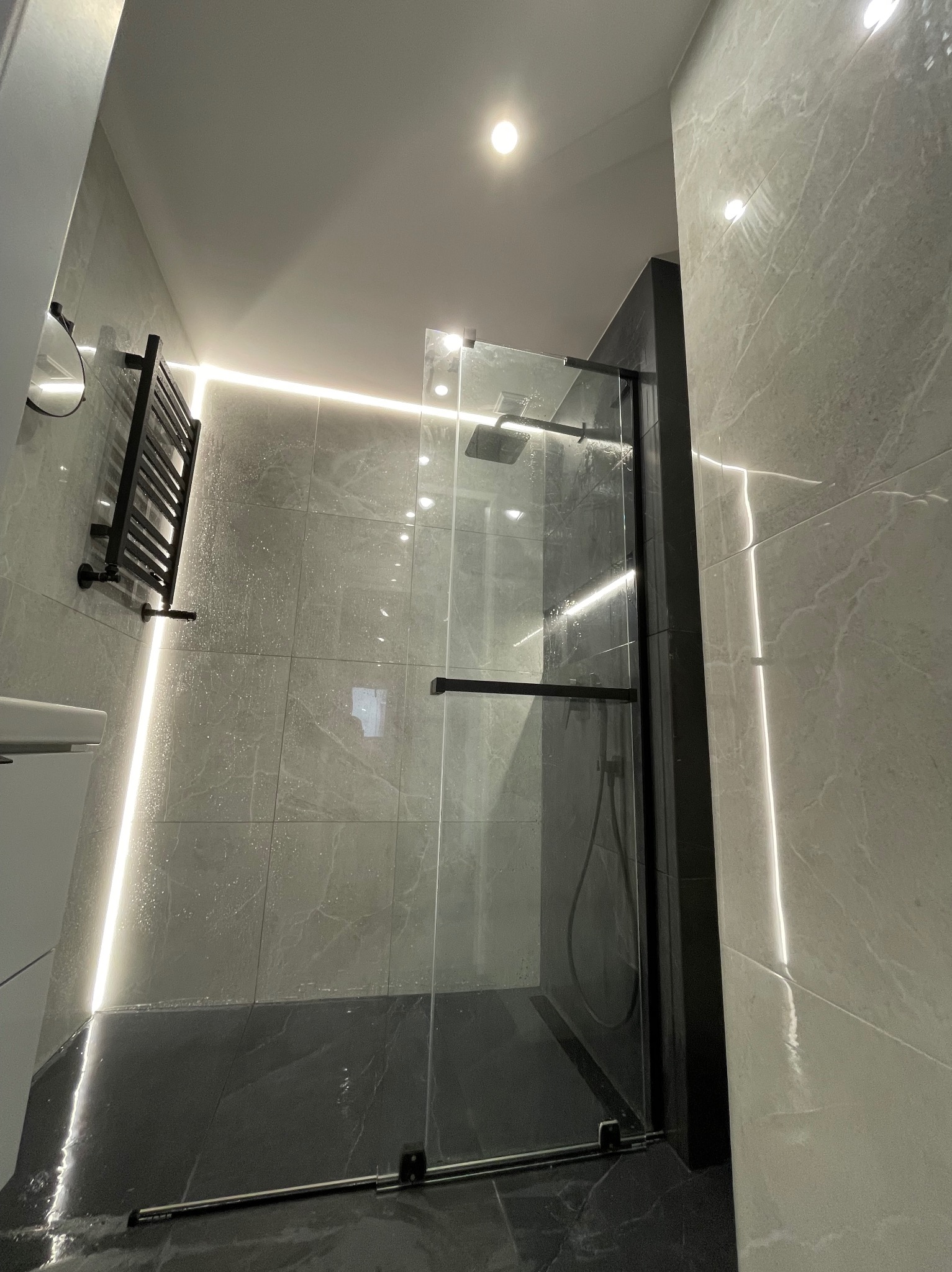 Nowoczesna łazienka po remoncie: szklana kabina prysznicowa z czarnymi ramami, marmurowe płytki ścienne i podłogowe, podświetlenie LED. Grzejnik drabinkowy.