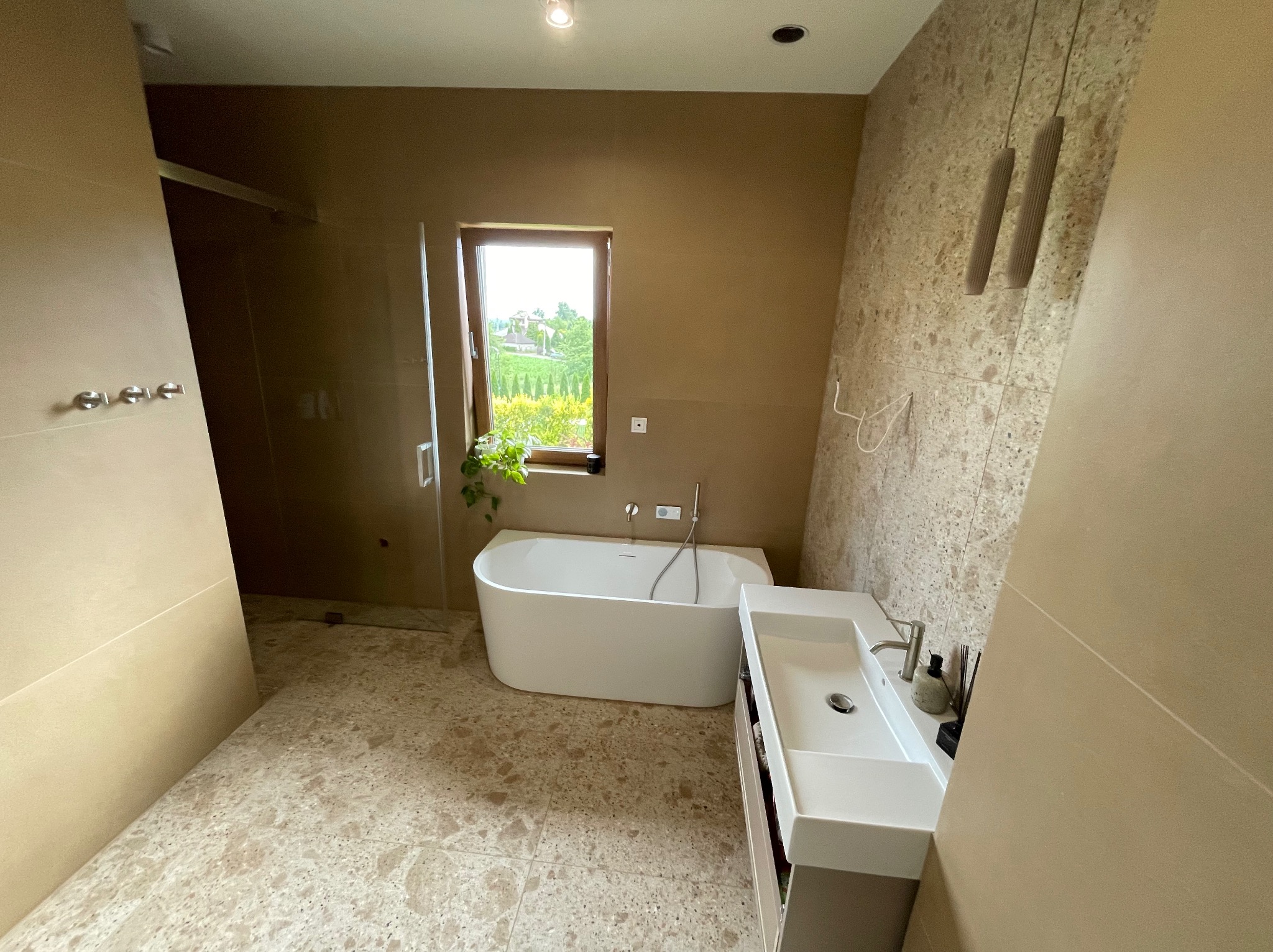 Nowoczesna łazienka w beżach: wolnostojąca wanna, umywalka z szafką, okno z widokiem na zieleń. Płytki podłogowe imitujące kamień, minimalistyczne oświetlenie.