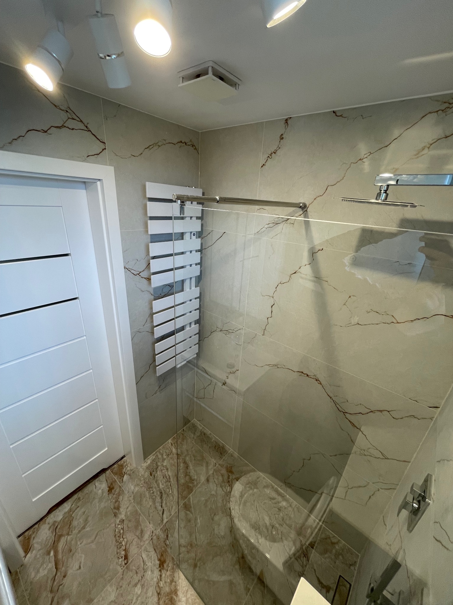 Nowoczesna kabina prysznicowa z marmurowymi płytkami, szklaną ścianką i grzejnikiem drabinkowym. Widoczne oświetlenie punktowe i białe drzwi. Eleganckie wykończenie.