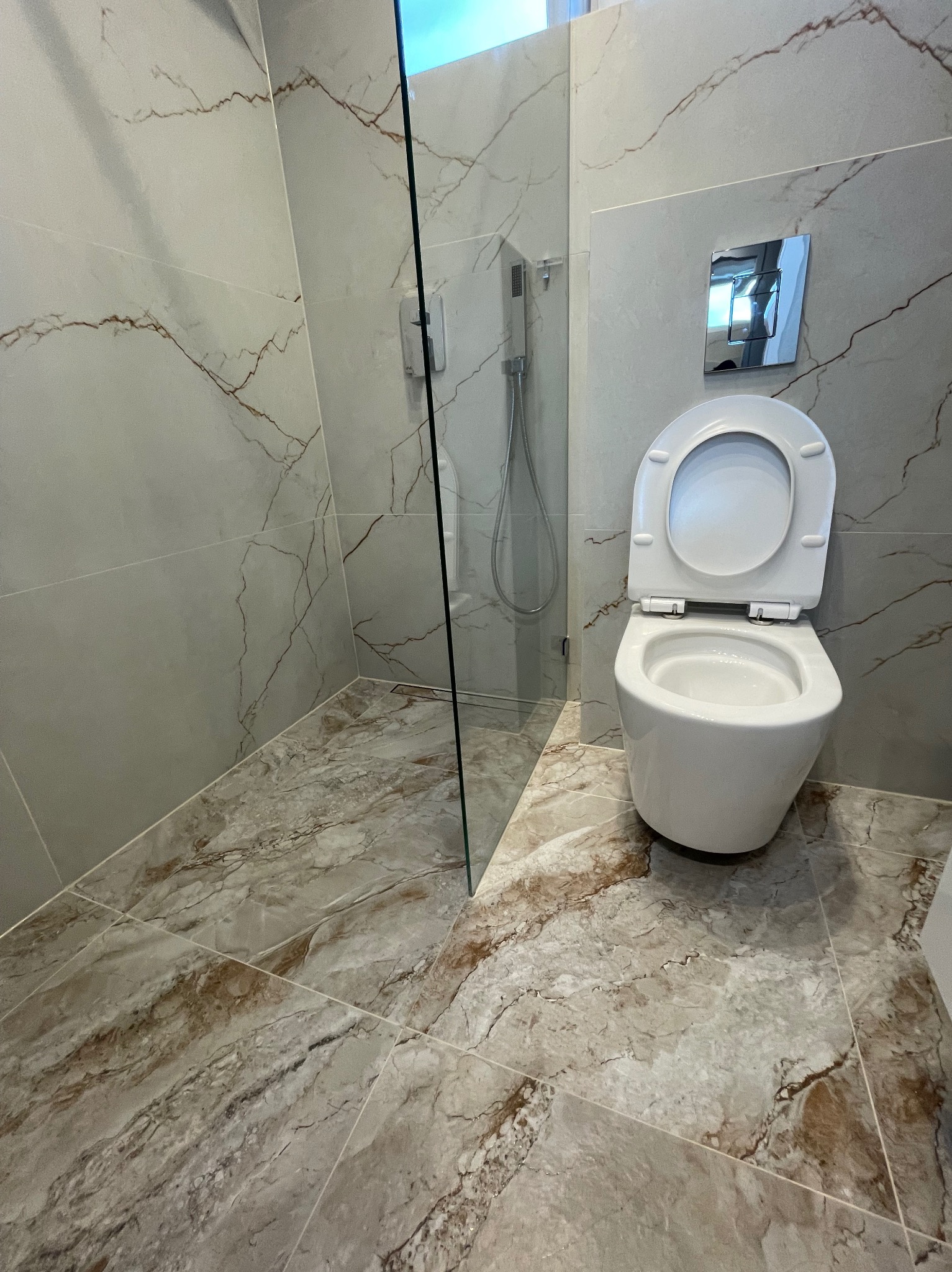 Wykończona łazienka z kabiną prysznicową typu walk-in i podwieszaną toaletą. Płytki imitujące marmur na ścianach i podłodze. Chromowana armatura i szklana ścianka.