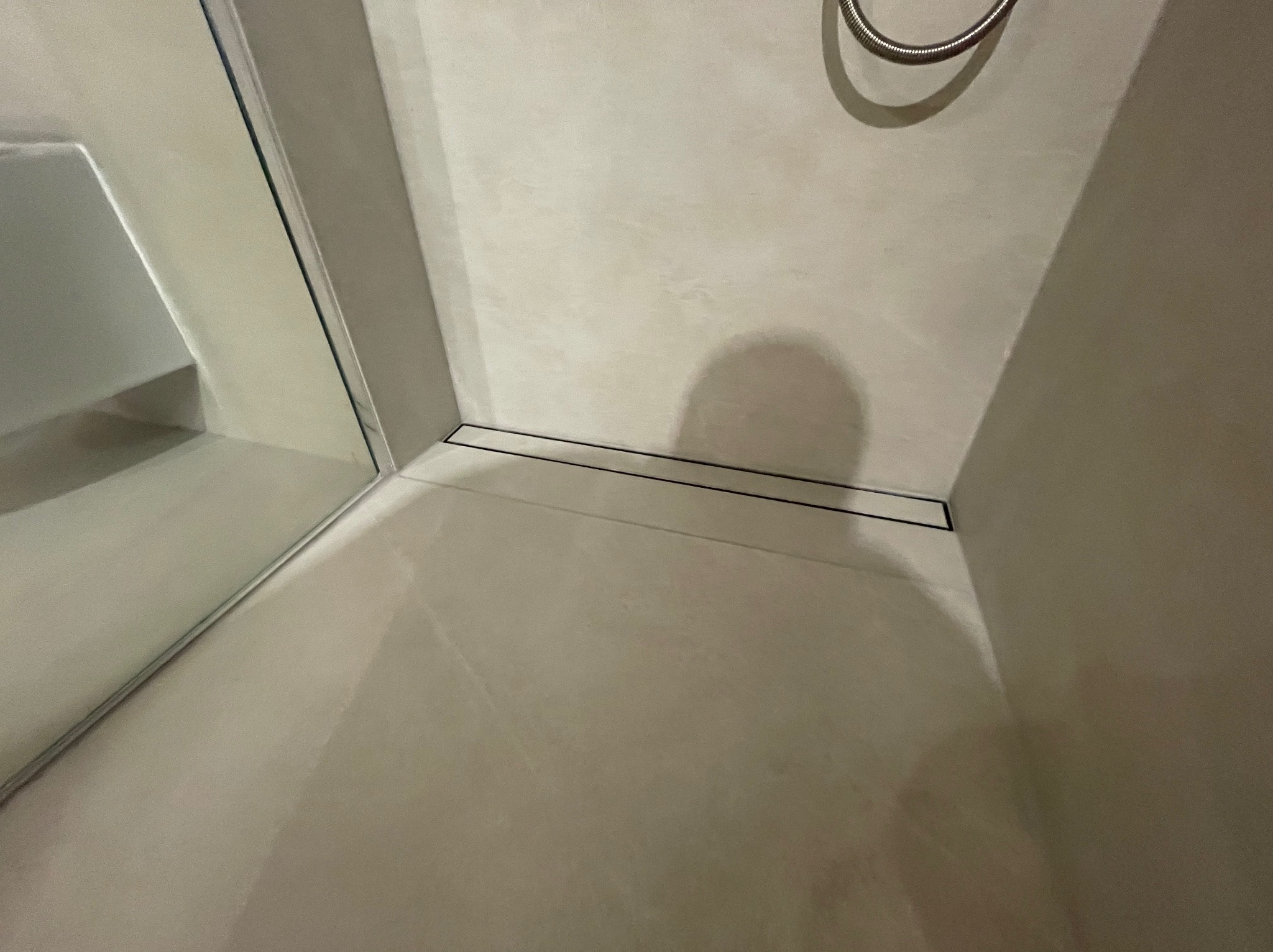 Minimalistyczna kabina prysznicowa z odpływem liniowym w podłodze, wykończona w jasnych, beżowych tonacjach. Widoczny fragment słuchawki prysznicowej.