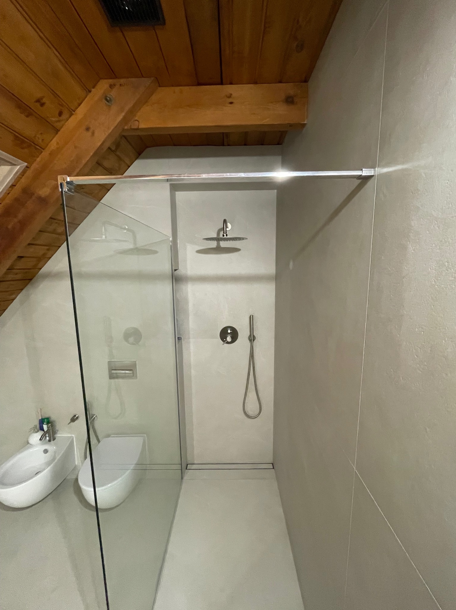 Nowoczesna łazienka z kabiną typu walk-in, bidetem i toaletą w minimalistycznym stylu. Uwagę przyciąga drewniany strop nad kabiną.