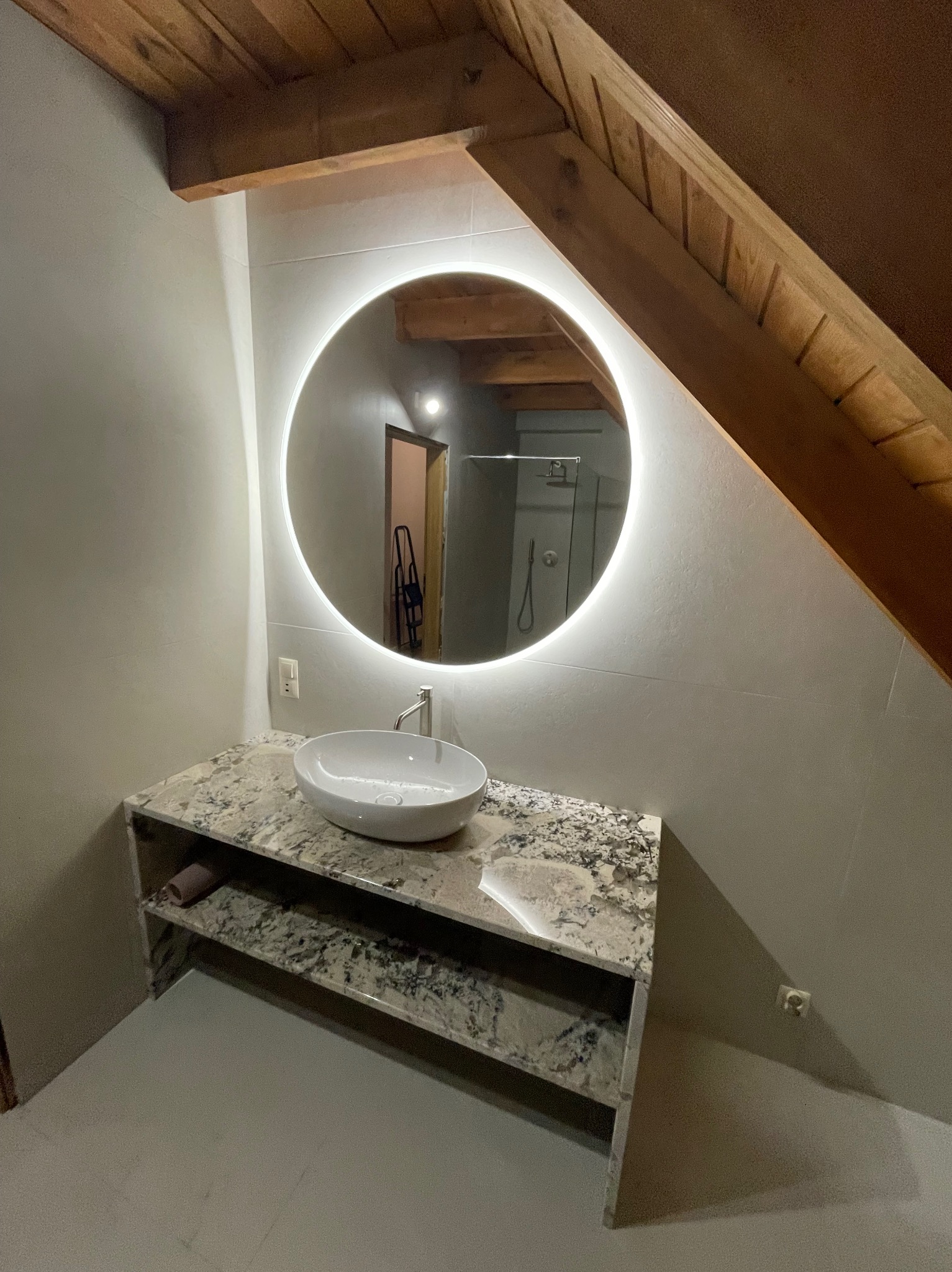 Wykończona łazienka na poddaszu: okrągłe lustro z podświetleniem LED nad umywalką z blatem imitującym marmur. Widoczne drewniane elementy konstrukcyjne dachu.