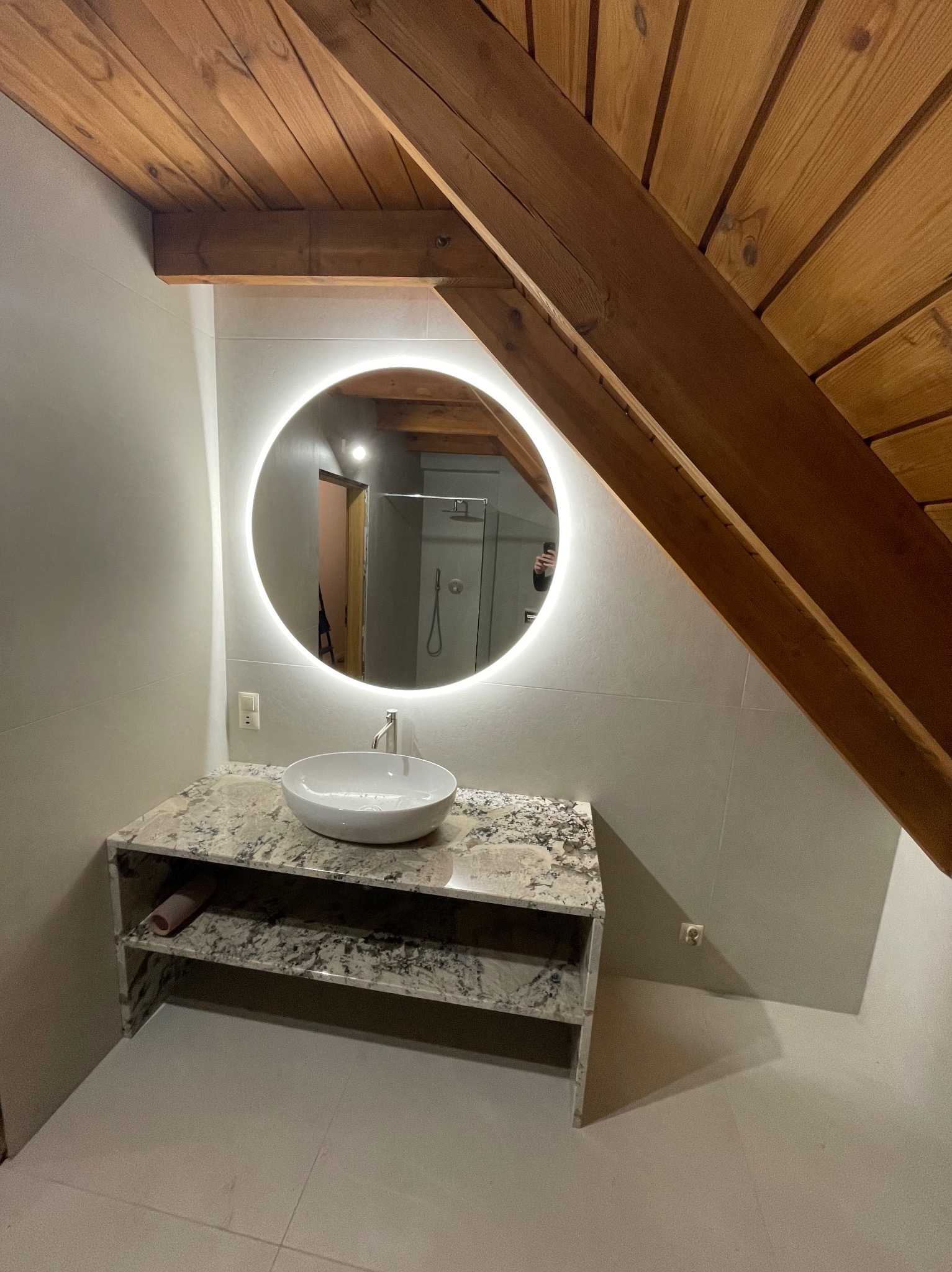 Nowoczesna łazienka na poddaszu z okrągłym lustrem LED, umywalką nablatową na blacie z kamienia i drewnianym stropem. Minimalistyczny design, jasne płytki.