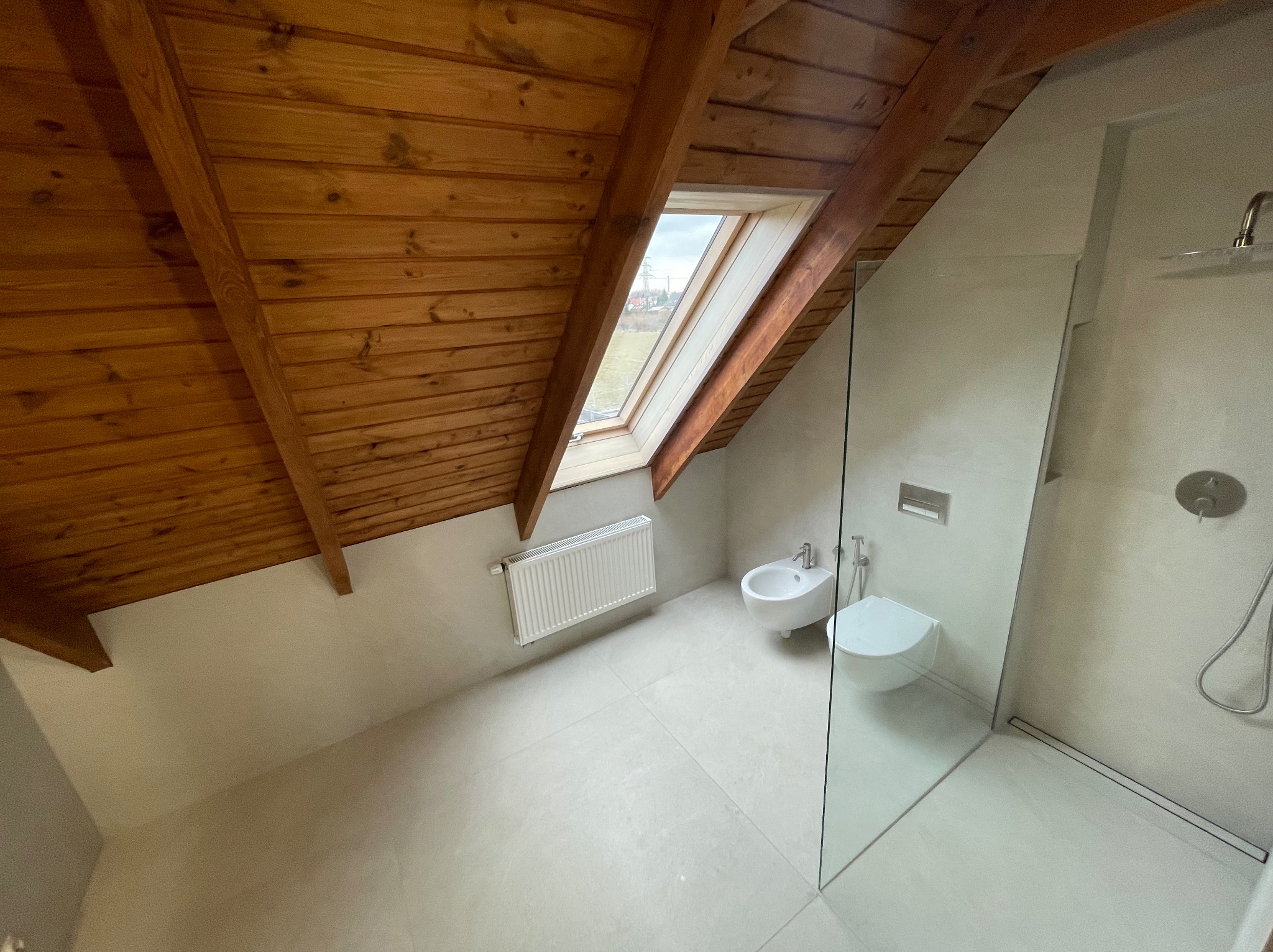 Jasna łazienka na poddaszu ze skośnym drewnianym sufitem i oknem dachowym. Kabina prysznicowa ze szklaną ścianą, toaleta, bidet i grzejnik.