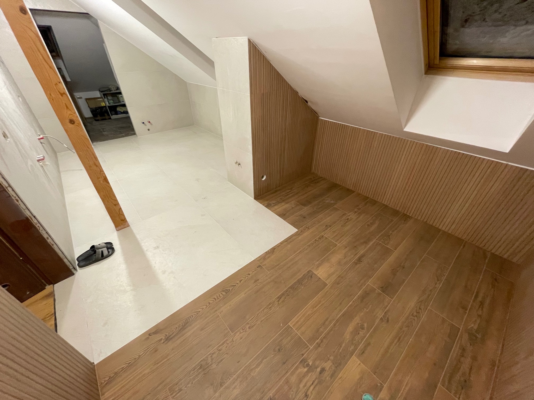 Wykończona łazienka na poddaszu z drewnopodobną glazurą na podłodze i ścianach, białe płytki na pozostałej części ścian. Widoczne skośne ściany i okno dachowe.