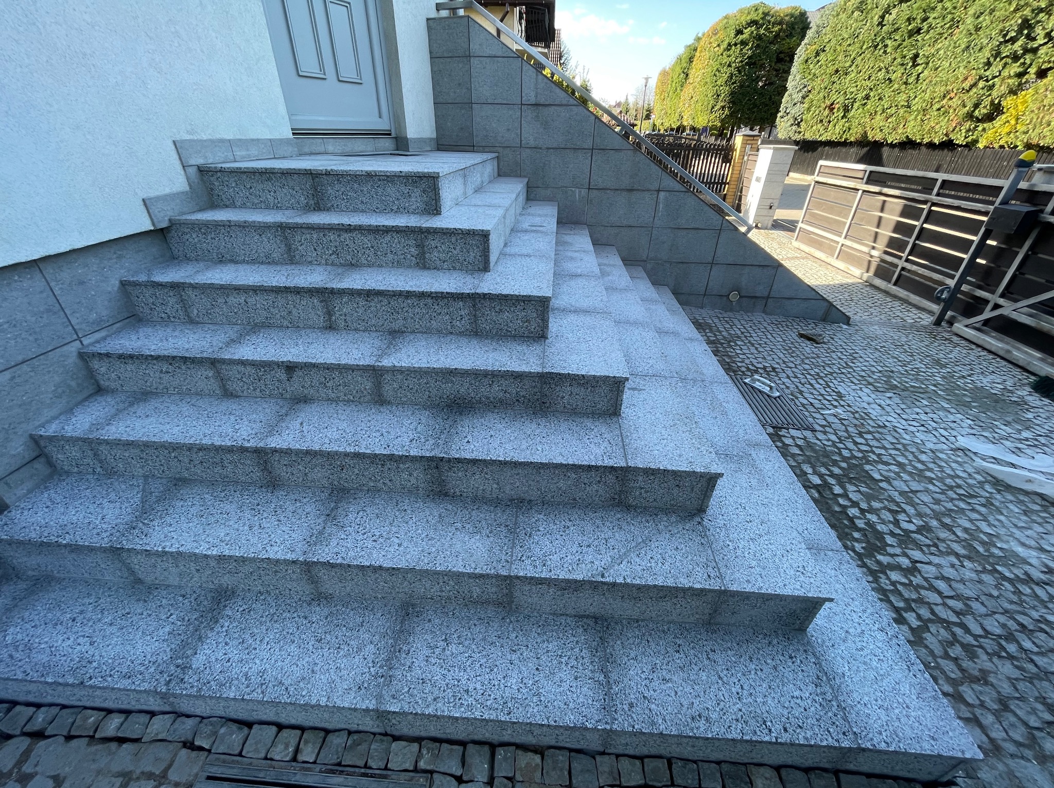 Granitowe schody wejściowe z metalową barierką, prowadzące do białych drzwi. Tło stanowią zielone krzewy i kostka brukowa.