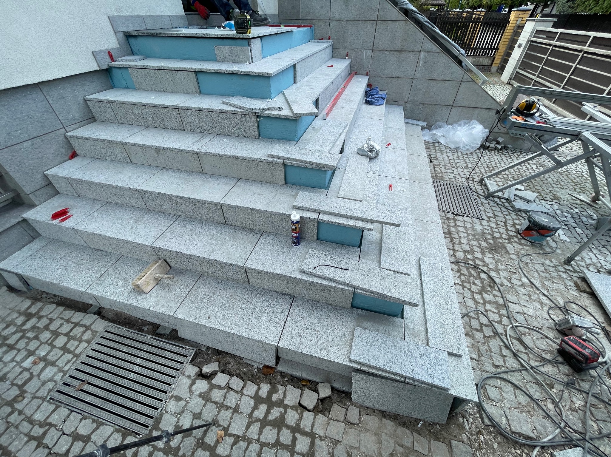 Montaż granitowych płytek na schodach zewnętrznych. Widoczne narzędzia glazurnicze, poziomica, klej. Etap prac: płytki częściowo ułożone, z niebieską izolacją.
